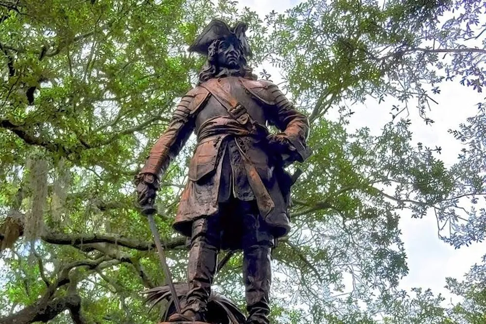 This photo depicts a bronze statue of a historical figure standing confidently with his left hand on his hip and his right hand holding a sword set against a backdrop of lush green trees