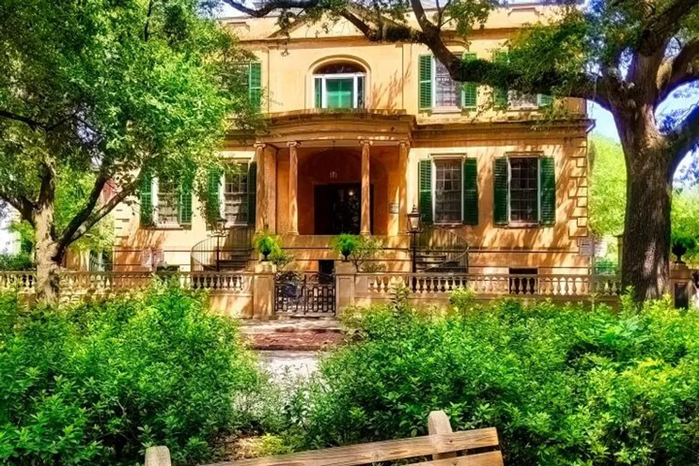 The image shows an elegant two-story house with a terracotta facade shuttered windows and a grand entrance nestled amongst lush greenery and mature trees under a sunny sky
