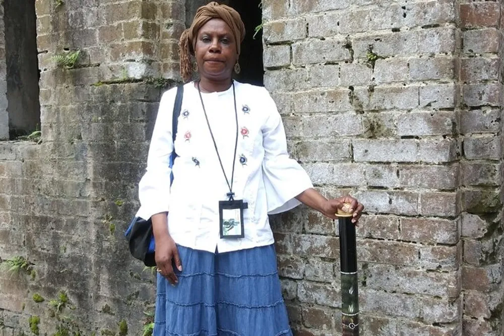 A woman is standing against a brick wall with her hand resting on an umbrella wearing a white blouse with floral detail a blue skirt and a badge around her neck