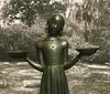 The image shows a bronze statue of a young girl holding two bowls out at her sides standing in front of a blurred backdrop of foliage