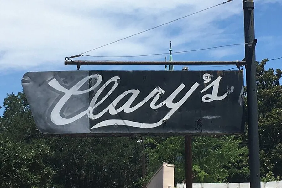 The image shows a weathered sign with the name Clary's in cursive script against a blue sky with trees in the background.