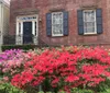 The image shows a traditional brick house with black shutters and a black front door complemented by a vibrant display of red and purple azalea bushes in bloom in the front yard