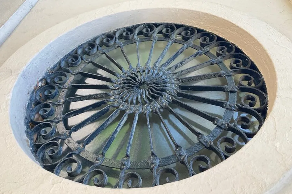 This is a photograph of an ornate circular wrought iron grate set within an arched alcove on a wall suggesting a decorative element or a vent for airflow