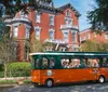 An orange and green trolley filled with passengers tours by a stately red brick Victorian-style building on a sunny day