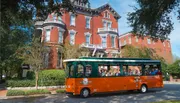 An orange and green trolley filled with passengers tours by a stately red brick Victorian-style building on a sunny day.