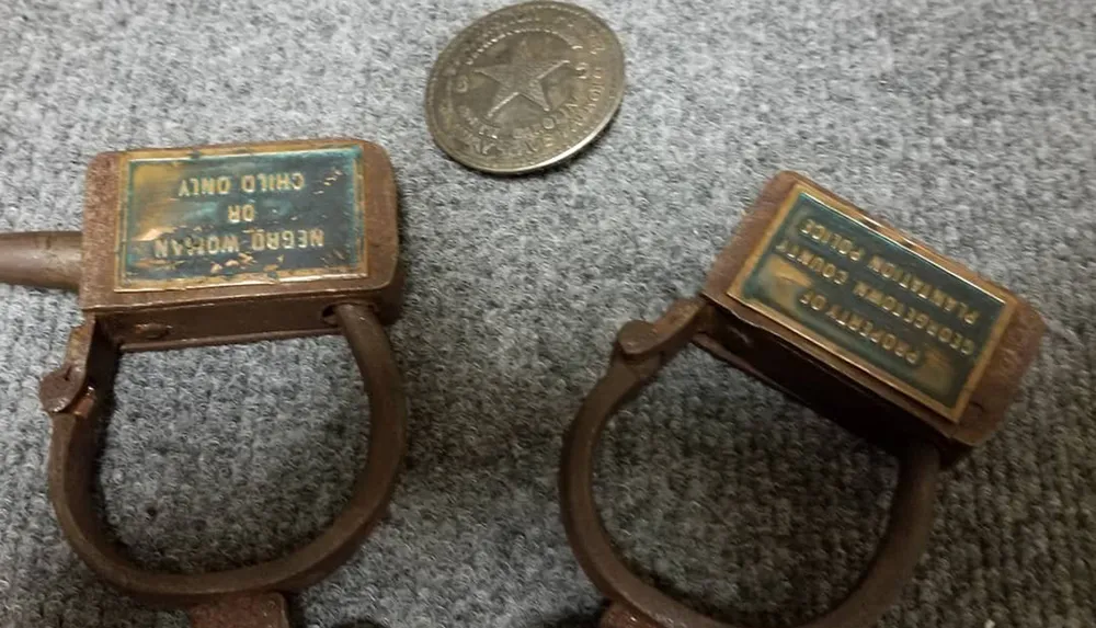 The image shows two antique-looking handcuffs with embossed brass plaques next to an old coin for scale all resting on a grey fabric surface