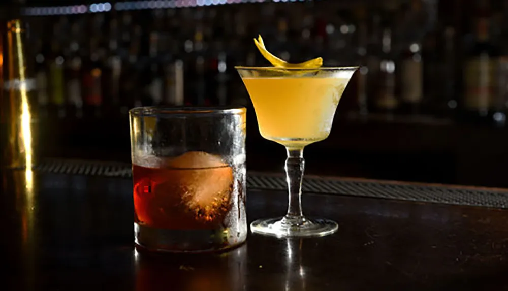 The image shows two cocktails on a bar counter one in a lowball glass with a reddish hue and ice and the other in a martini glass with a yellowish color and a lemon twist garnish