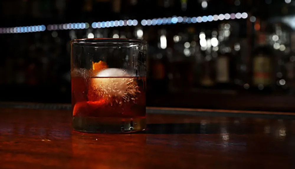 There is a glass containing a dark-colored beverage with a large ice cube and an orange peel on a bar with bottles in the softly lit background