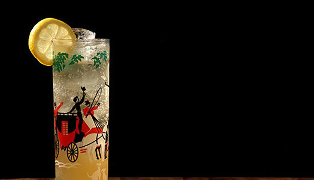The image shows a refreshing drink with a slice of lemon on the rim served in a tall glass with a decorative print against a dark background