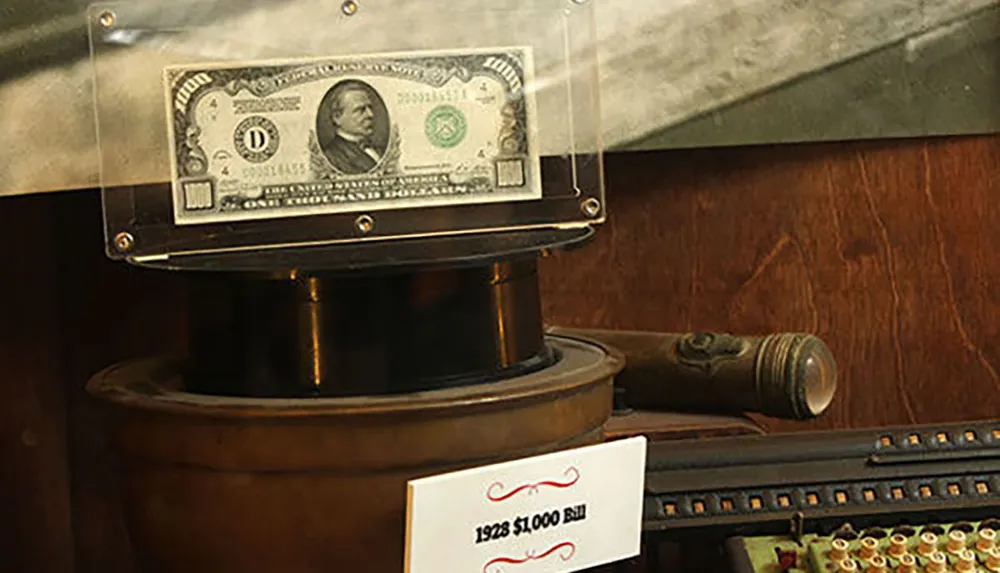 The image shows a 1928 1000 bill on display protected inside a clear case mounted on a pedestal with a sign indicating the bills denomination and year and set against a backdrop of vintage items