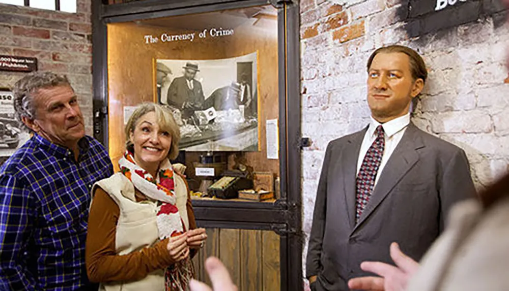 A man and a woman are posing with a lifelike figure inside a room that appears to be part of an exhibition titled The Currency of Crime