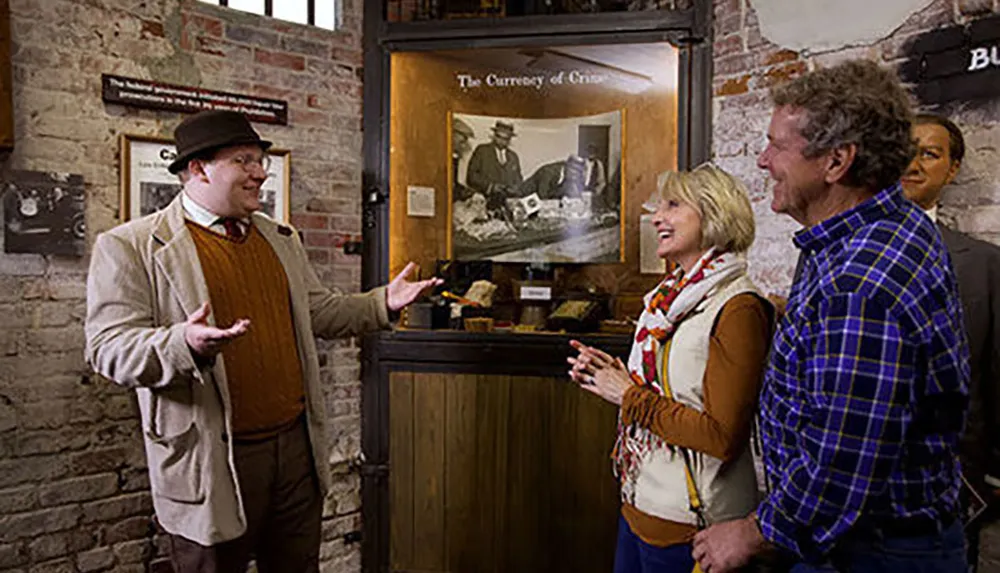 A man wearing a hat and glasses is standing in front of an exhibit titled The Currency of Crisis gesturing while explaining something to an engaged couple who are smiling and listening intently