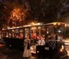 A trolley bus with passengers is parked at night under tree-lined streets accompanied by two individuals in period clothing by its side
