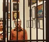 A man and a woman stand behind a set of vertical bars with various framed artworks on the wall behind them