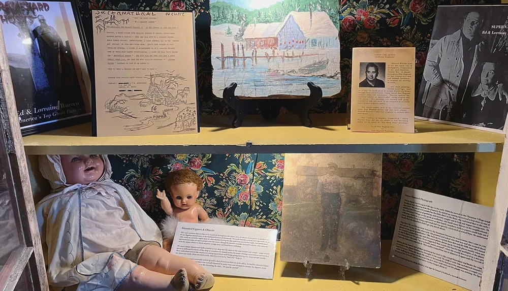 The image shows a display case with various items including old dolls framed pictures and possibly historical documents potentially part of a museum exhibit or collection related to local history or folklore