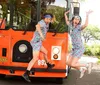 Two individuals wearing matching tropical-themed clothing and headbands pose humorously in front of a trolley promoting a tour named Savannah for Morons