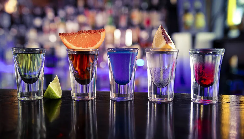 Five colorful shot glasses each garnished differently are lined up on a bar counter with a reflective surface and a blurred background of illuminated liquor bottles