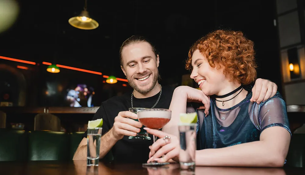 A man and a woman are sharing a cheerful moment over drinks at a bar with warm lighting in the background