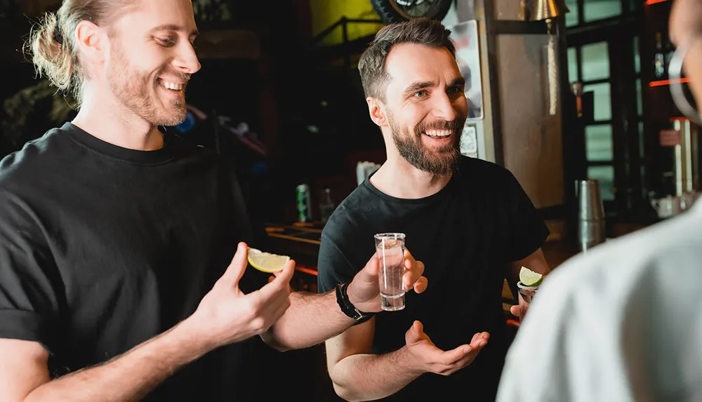 Two men are smiling and holding slices of lime and a shot glass appearing to be enjoying a social moment at a bar