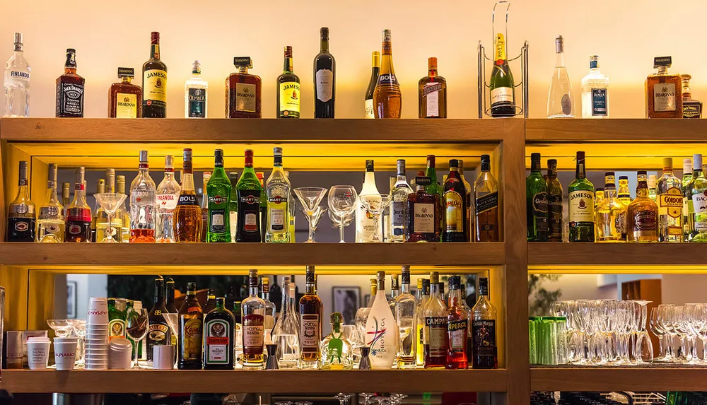 The image shows a well-stocked bar shelf with an array of various liquor bottles and glassware under warm lighting
