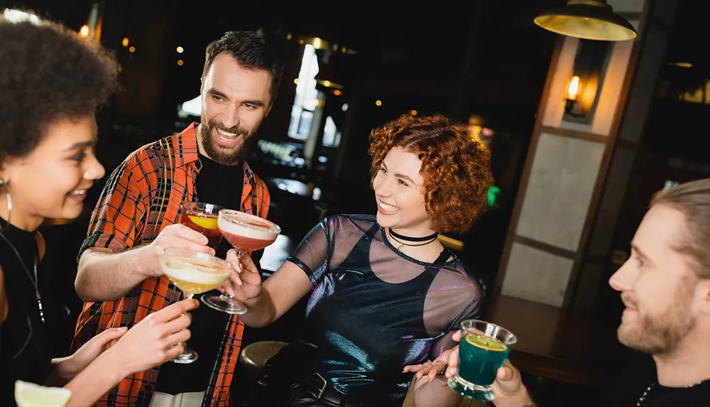 Four friends are cheerfully toasting with colorful cocktails in a cozy bar setting