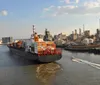 A large cargo ship loaded with containers is navigating a river near industrial facilities while a small boat speeds by in the foreground