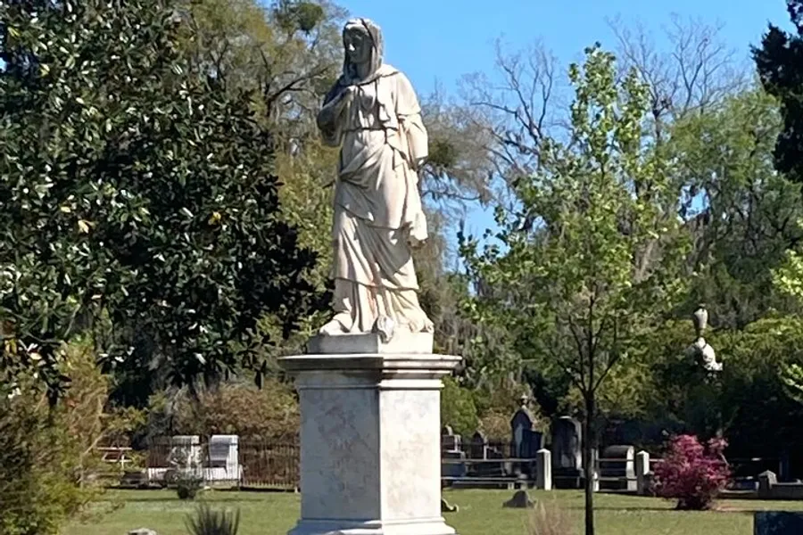 The image shows a white marble statue of a draped figure, likely representing a classical or allegorical figure, situated outdoors in a serene cemetery surrounded by trees and other monuments.