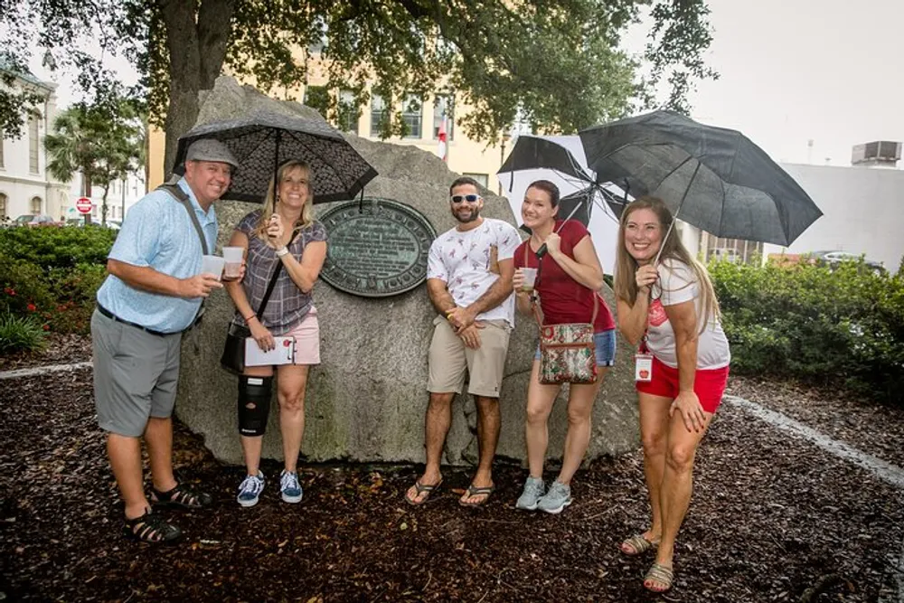 A group of people are smiling and posing with umbrellas next to a historical marker on a cloudy day