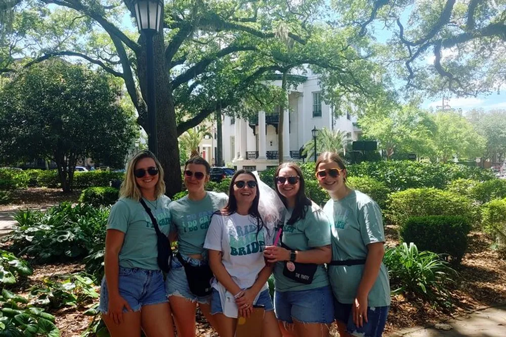 Five people are posing for a photo in a sunny park-like setting with trees and a white building in the background and some of them are wearing matching green t-shirts