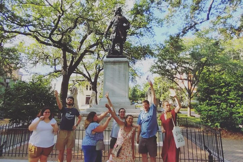 A group of people is posing joyfully in front of a statue in a park with lush green trees in the background