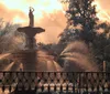 The image shows an ornate water fountain with spraying water backlit by a glowing sunset surrounded by trees and enclosed by an elegant fence