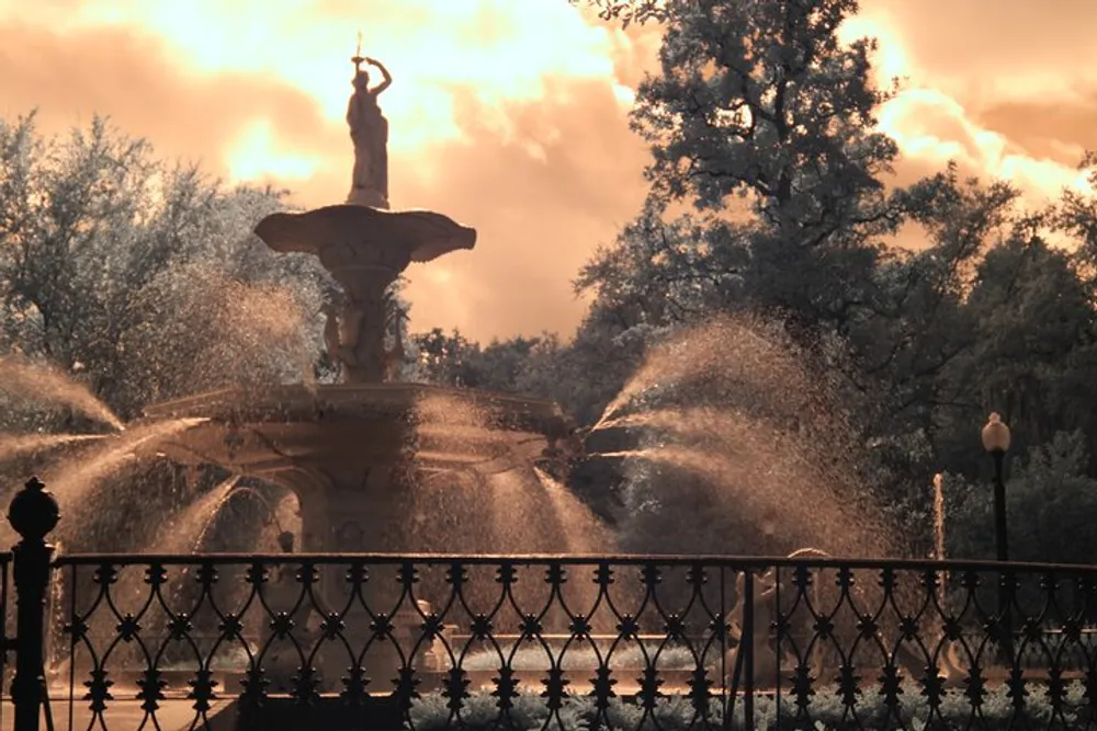 The image shows an ornate water fountain with spraying water backlit by a glowing sunset surrounded by trees and enclosed by an elegant fence