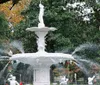 An ornate white fountain with water spouts adorned with festive red ribbons is surrounded by lush greenery and a lamppost