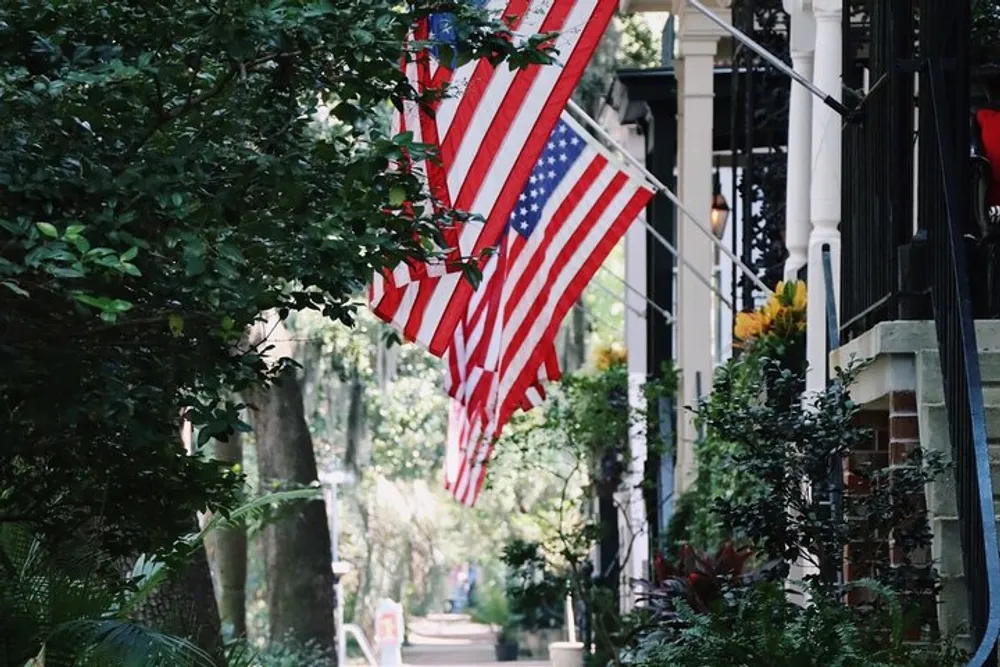 Several American flags are hanging from the porches of a quaint street lined with greenery and historic buildings