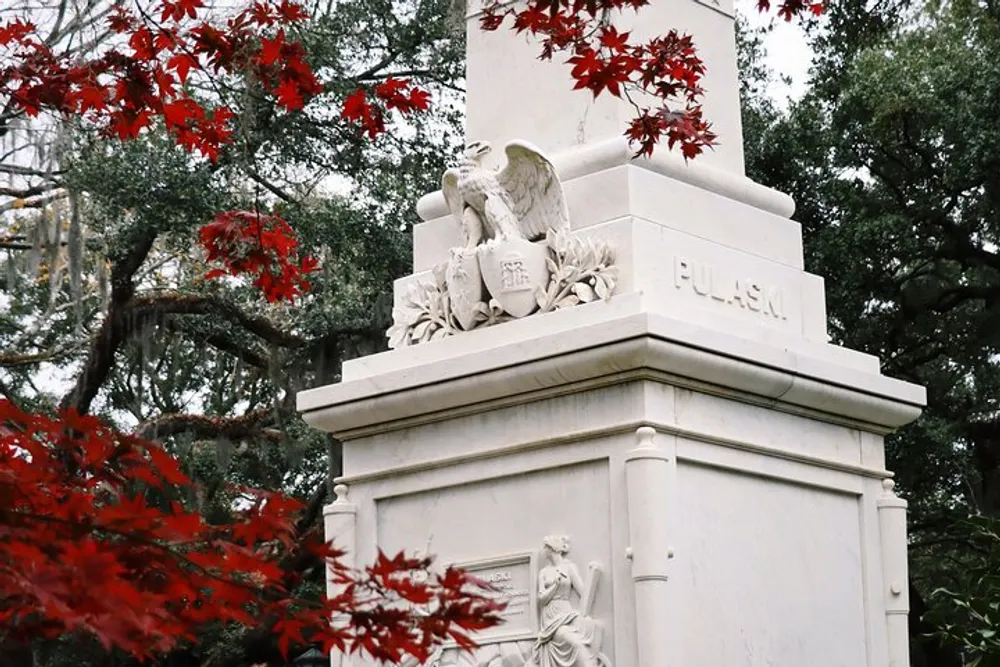 The image shows a white monument with the name PULASKI at the top adorned with sculptural elements framed by vibrant red leaves in the foreground and green foliage in the background