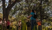 A group of people listen attentively to a speaker outdoors on a sunny day, surrounded by lush greenery.