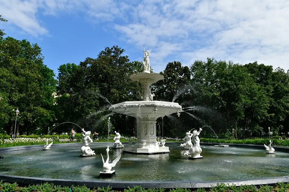 An elaborate white fountain with multiple tiers and sculptural figures is surrounded by verdant greenery under a clear blue sky