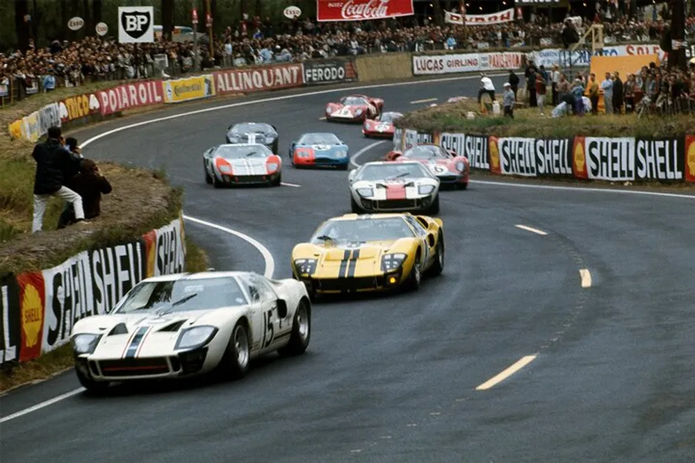 Vintage race cars including a prominent Ford GT40 are competing on a curvy track surrounded by spectators and vintage advertising banners