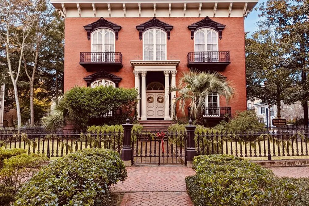 The image shows a symmetrical red-brick house with black wrought iron railings two levels of ornate black balconies a grand entrance and a manicured garden evoking a sense of historic elegance