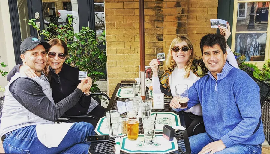 Four people are smiling and holding up cards while sitting at a restaurant table outdoors.
