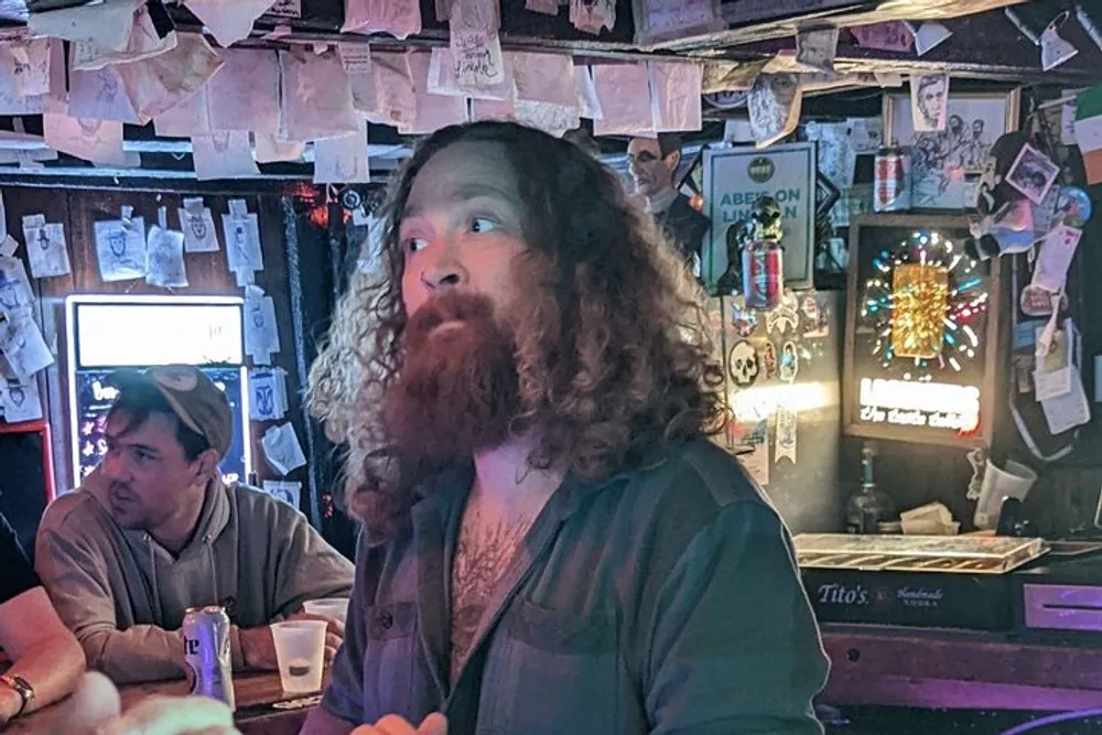 A man with curly hair and a beard is looking away from the camera with an interested expression inside a bar decorated with dollar bills and various memorabilia