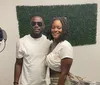 A man and a woman are smiling for a photo in a room with music equipment and a wall decorated with artificial greenery