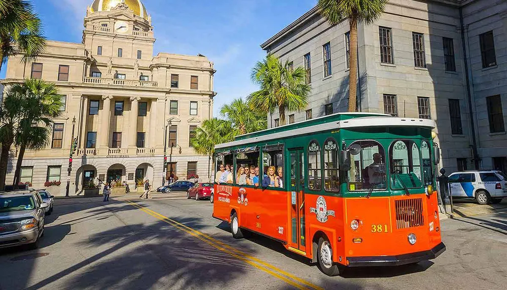 A vibrant orange and green trolley bus filled with passengers tours through a sunny palm-lined street in front of an ornate building with a golden dome