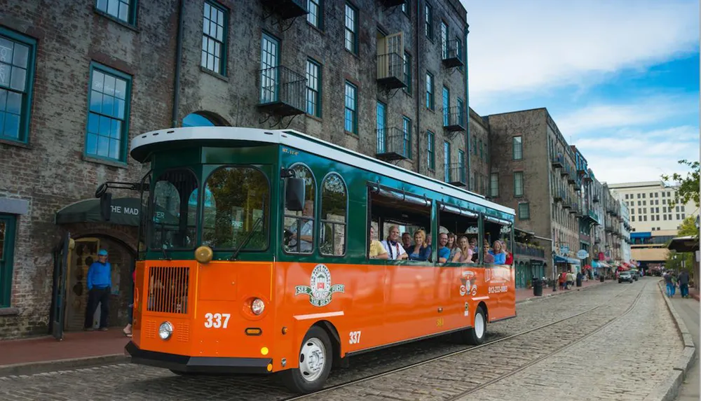A trolley full of passengers tours down a street lined with historic-looking buildings with cobblestone pavement and a railway track