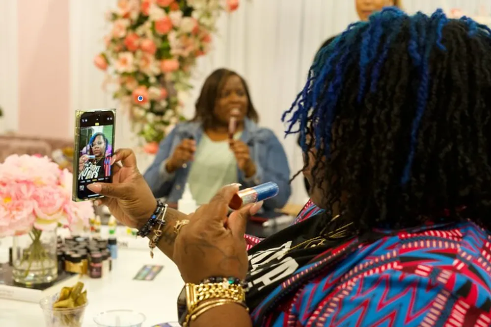 A person with blue braided hair is taking a selfie with a smartphone while another individual speaks into a microphone in the background at a floral-decorated event