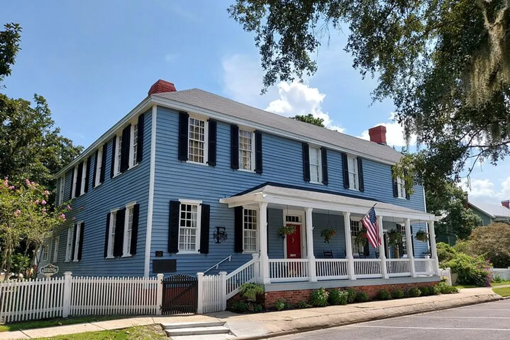 A classic two-story blue house with white trim a red front door an American flag and a welcoming front porch under a sunny sky
