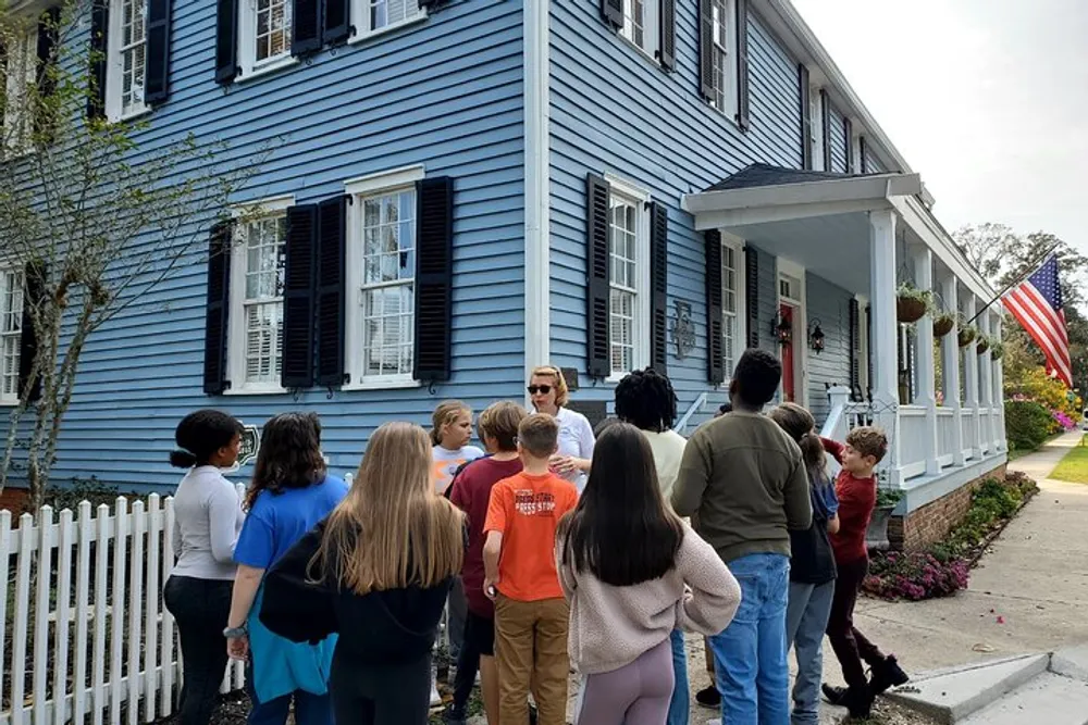 A group of people is gathered outside a blue house with black shutters listening to a person presumably giving a tour or presentation
