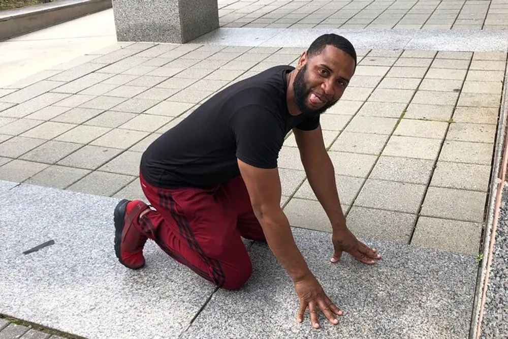 A man is crouching on one knee on the pavement smiling towards the camera with a playful expression