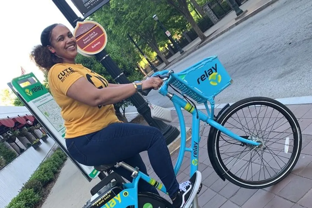A person is smiling while sitting on a blue and yellow Relay bike rental in an urban setting