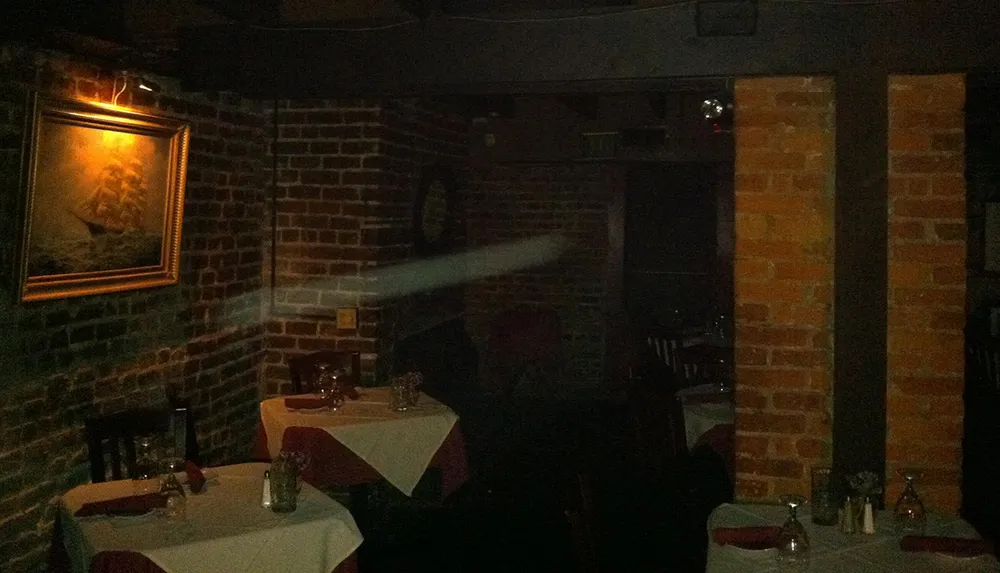 This image portrays a dimly lit seemingly empty restaurant with brick walls framed artwork and tables set for dining